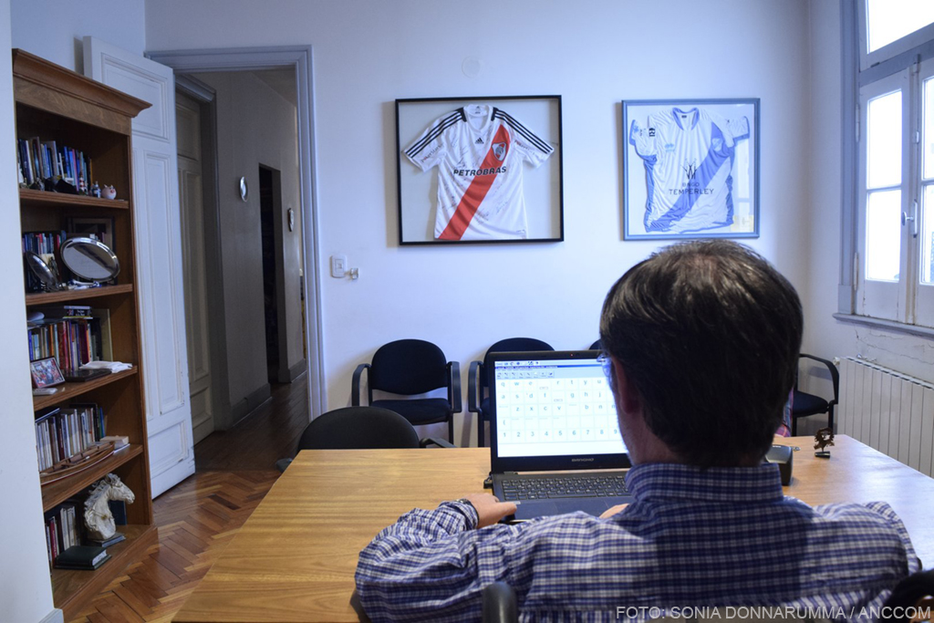 Se ve la espalda de Rivas quien trabaja con la computadora en su oficina. En la pared que tiene de frente se pueden observar dos camisetas de River enmarcadas y colgadas. 