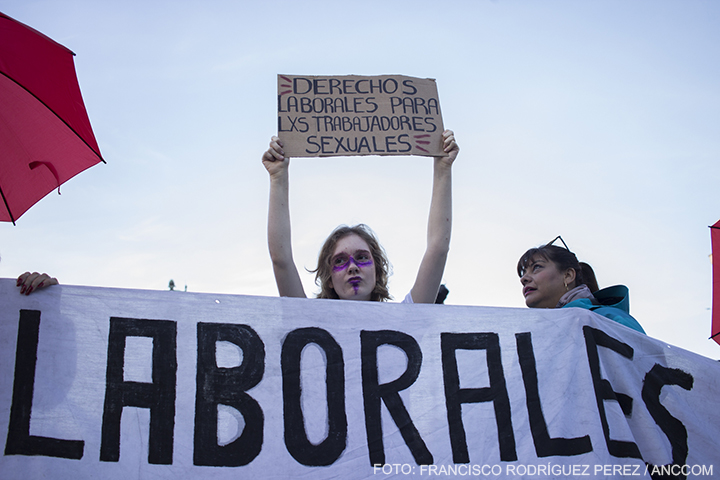 Mujera con la cara pintada levanta un cartel que dice "DERECHOS LABORALES PARA LXS TRABAJADORES SEXUALES"