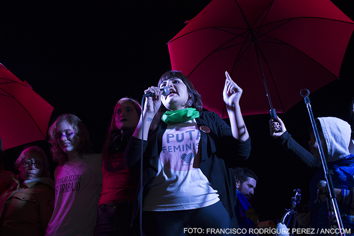 Una mujer hablando por micrófono en el escenario, lleva la frase "PUTA FEMINISTA" en su remera. De atrás se observan las demás oradoras del acto, y unos paraguas rojos, símbolos del trabajo sexual.
