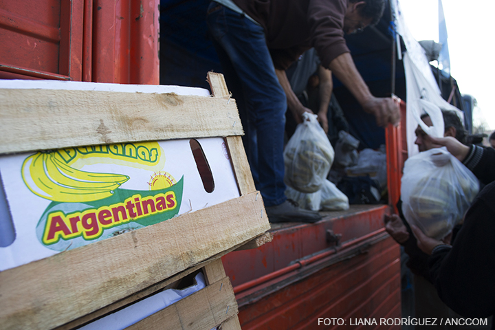 En primer plano un cajón de bananas en el que se lee: "bananas argentinas" y en segundo plano se ven las manos de hombres repartiendo bolsas de bananas arriba de un camión.