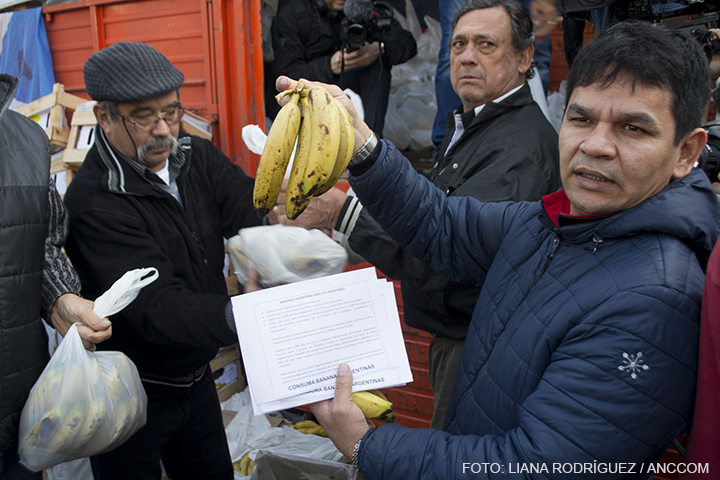 En primer plano se ve a un hombre sosteniendo con su mano en alto un manojo de bananas y con su otra mano sostiene un papel en el que se lee: "consuma bananas argentinas" 