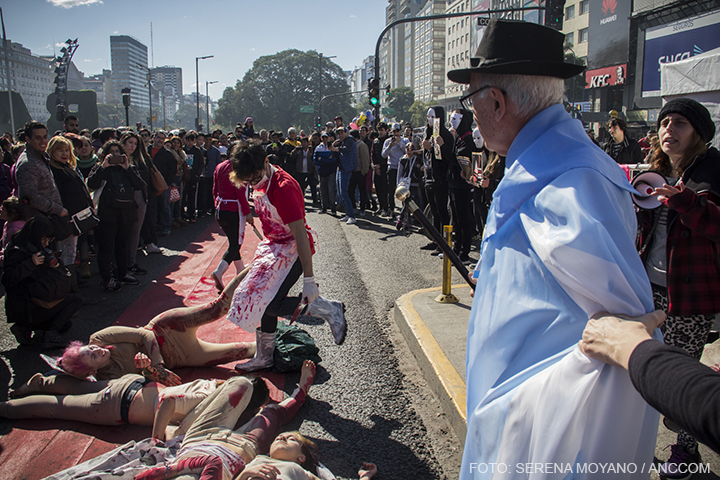 Un hombre mayor con un sombrero y envuelto en una bandera de Argentina es sostenido de un brazo mientras intenta acercarse a un grupo de activistas que se encuentran en el piso representando un matadero.