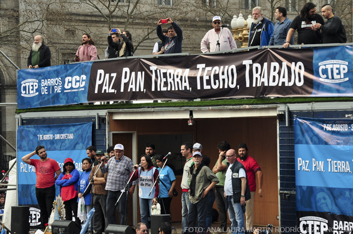 Un grupo de representantes de las organizaciones que convocaron la marcha se encuentran hablando desde un escenario, en una bandera que se encuentra encima del escenario se lee: "Paz, Pan, Tierra, Techo, Trabajo".