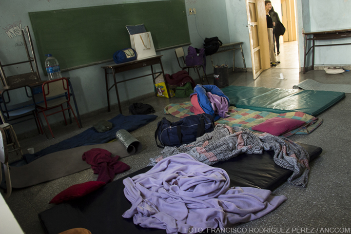 Un aula de colegio dentro de la misma, en el piso, se encuentran bolsas de dormir, mochilas, colchonetas y frazadas.