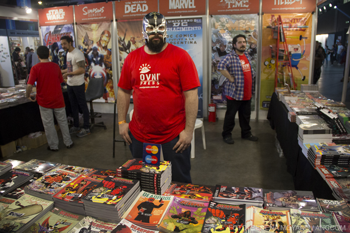 Un hombre usando una máscara se encuentra dentro de un stand, delante de él una mesa con una gran cantidad de historietas apiladas.