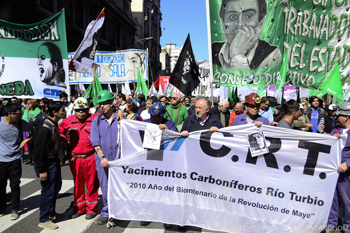 Gente marchando con distintas banderas verdes y blancas, delante una fila de hombres marchando vestidos con mamelucos y cascos con linternas sostienen una bandera en la que se lee: Yacimientos Carboníferos Río Turbio.