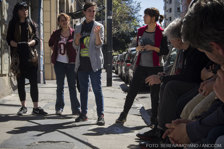 Se ve a un grupo de personas sentadas en la vereda de la calle y a tres mujeres paradas, una de ellas hablando con su mano levantada.