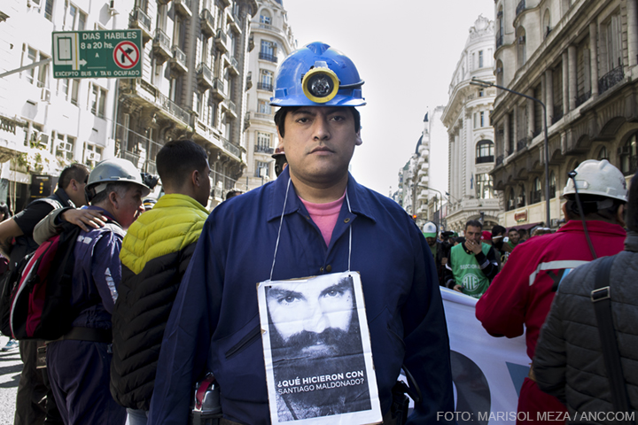 Un Hombre con ropa de minero y casco con linterna mira a cámara, de su cuello cuelga un cartel en el que se ve la imágen de Santiago Maldonado.