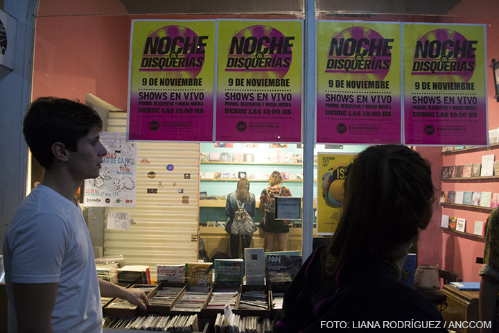 Una mesa repleta de CDs afuera de una disquería, dos jóvenes caminando, y en la vidriera unos afiches que promocionan "La noche de las disquerías".
