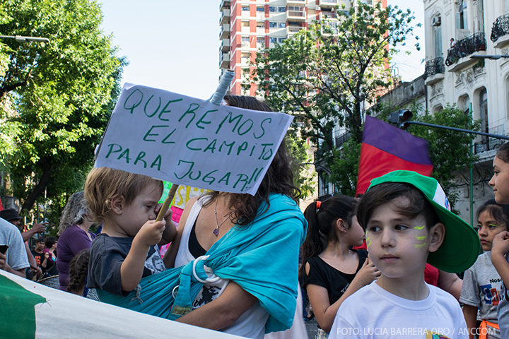 De la muchedumbre de la Caravana Artística, se destaca la presencia de dos niños. Uno sostiene un cartel que dice: "Queremos el campito para jugar".