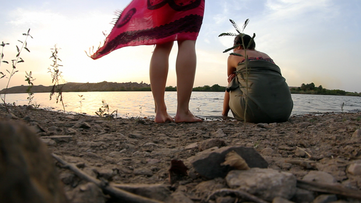 Imagen del documental "Wanderlust, cuerpos en tránsito". En una playa, dos mujeres, una sentada y la ota parada (solo seven sus piernas), mirando al mar.