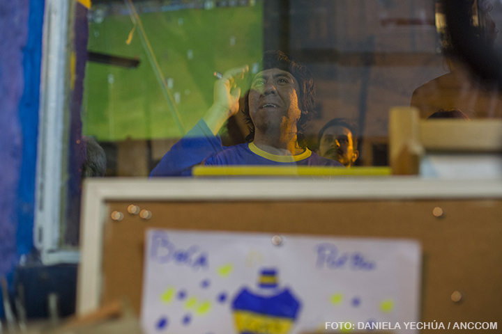 Detrás del vidrio del local de la Asociación Civil Boca es Pueblo, se puede observar a un hombre vestido con la camiseta de Boca que lamenta una situación. El reflejo en el vidrio devuelve la imagen difusa del televisor que transmite el partido.