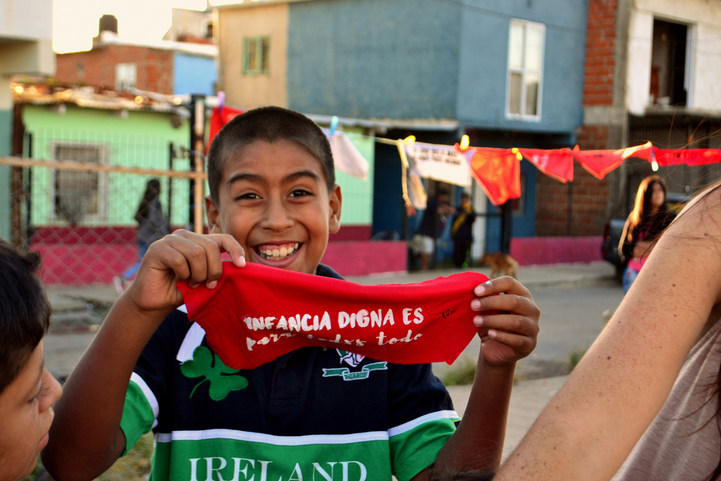 Imagen de un niño sonriendo y sosteniendo un pañuelo rojo que dice "infancia digna es..."