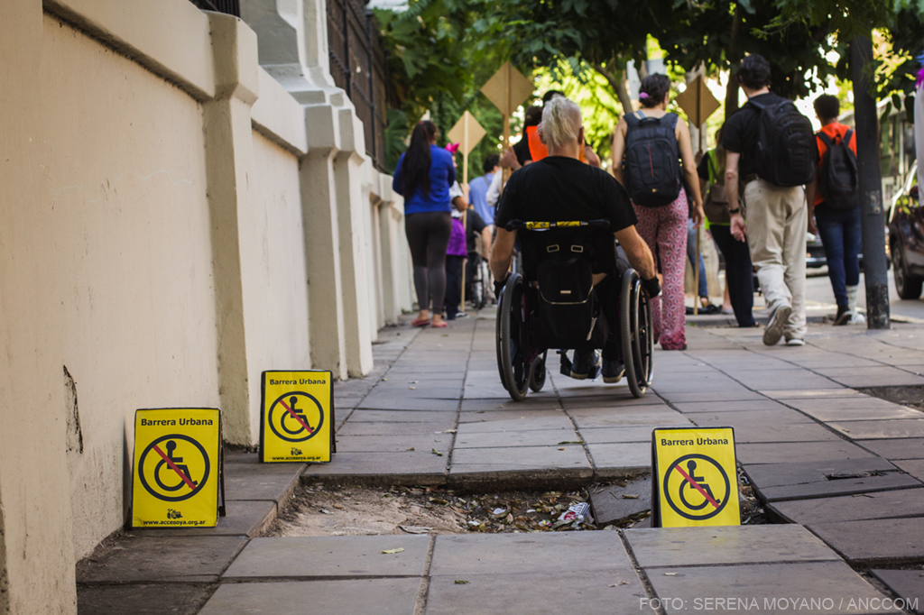 imagen de una vereda rota, en el fondo se ve la gente que realizó el recorrido caminando y en sillas de ruedas.
