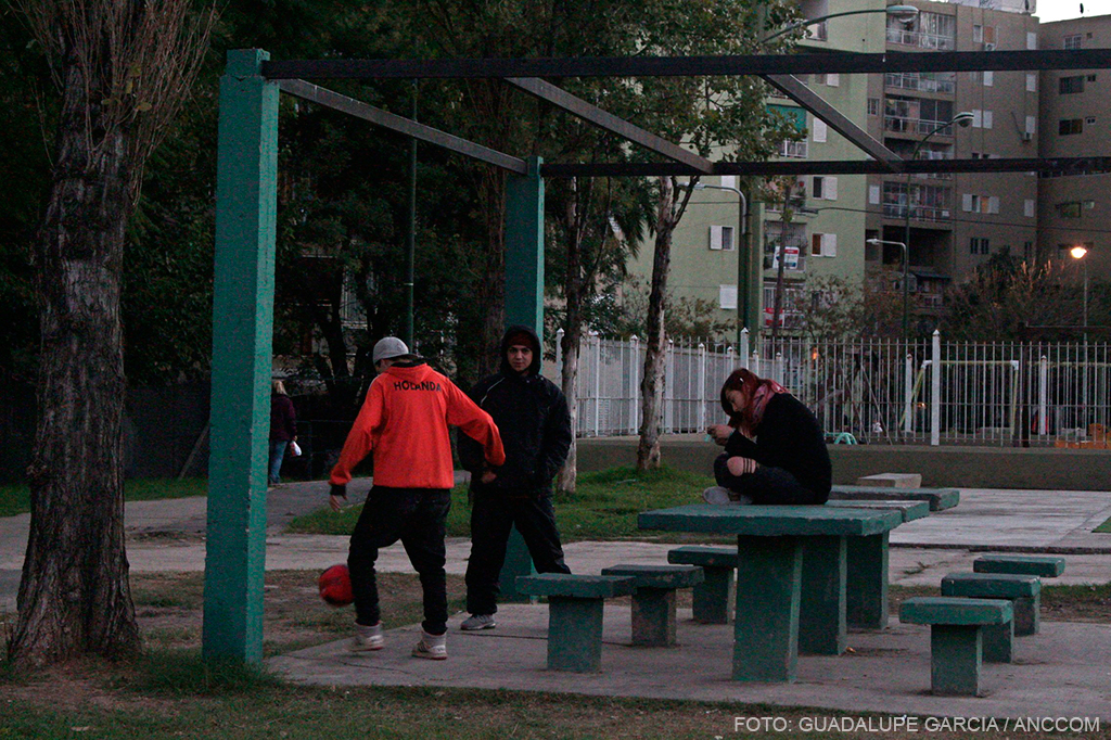 Tres adolescentes, dos chicos jugando con una pelota y una chica sentada sobre una mesa de cemento. De fondo se observan los monoblocks.