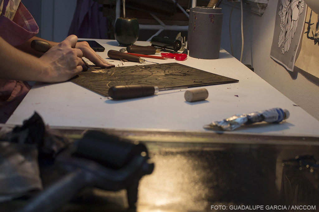 Manos tallando madera sobre un escritorio sobre el se observn objetos artísticos y un mate.
