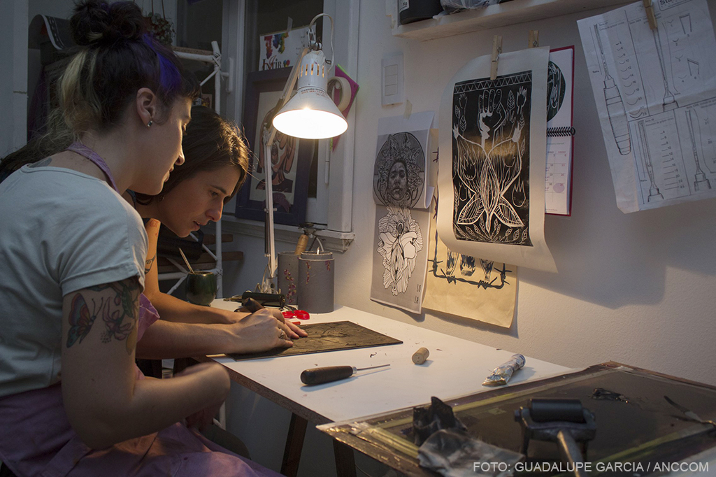 Dos mujeres tallando madera sobre un escritorio, se observan alrededor afiches colgados.