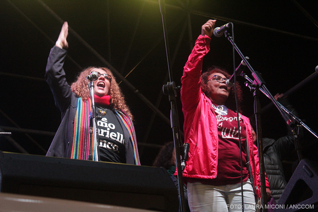 Dos mujeres con sus brazos en alto hablando sobre el escenario.