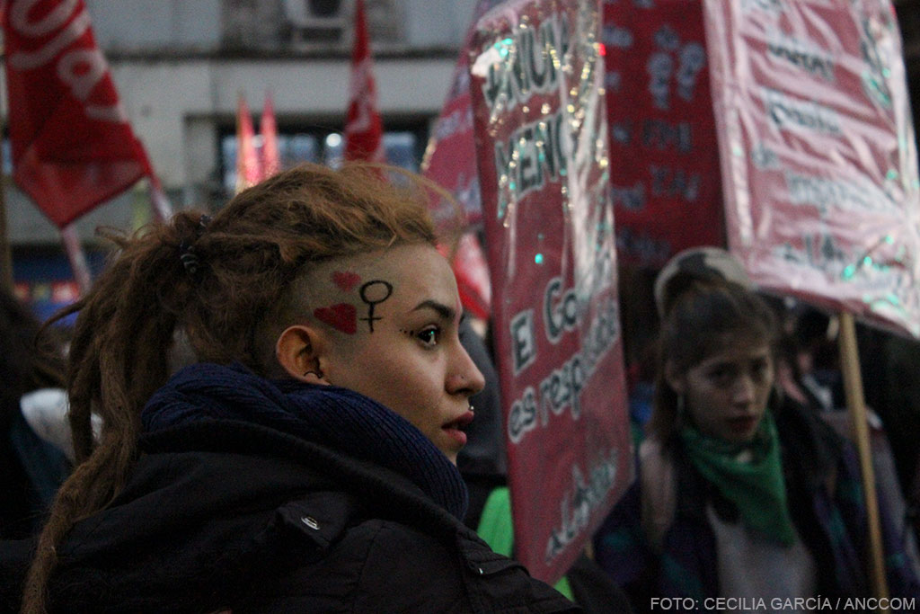 Mujer de perfil con rastas y su cara pintada, de fondo se observan otras mujeres y carteles rojos.