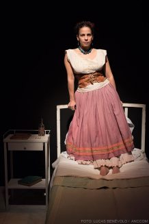 Actriz interpretando a Frida parada sobre una cama junto a una mesa de luz.
