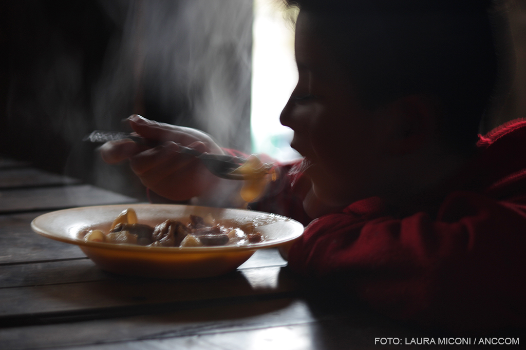 Figura de perfil de un niño llevándose a la boca un tenedor con comida. Frente a un plato de fideos humeando